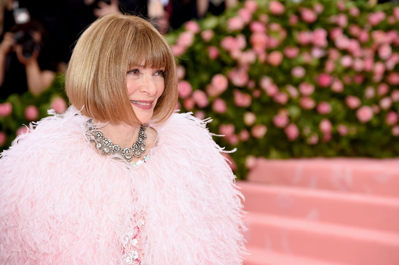 A woman with a blonde bob in a fluffy pink coat and a huge diamond necklace.