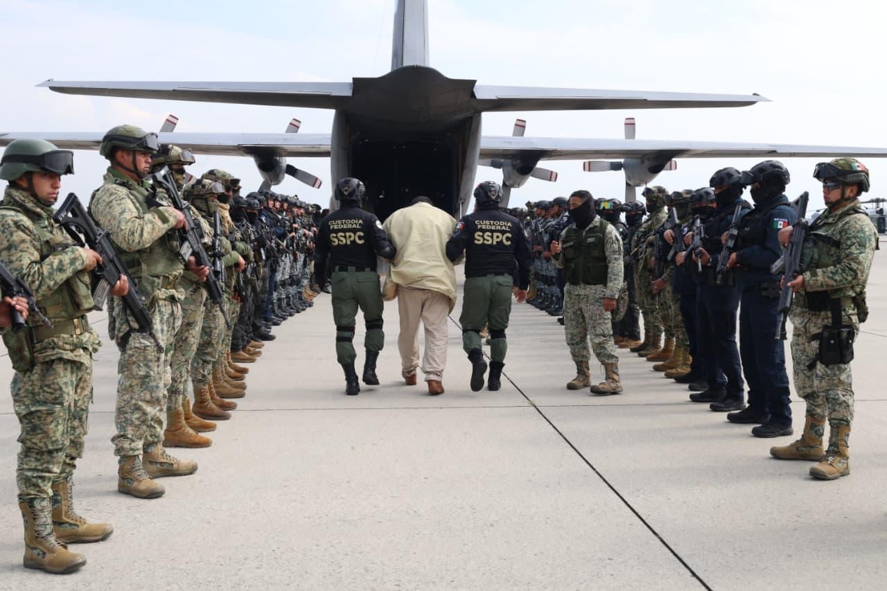 A man is escorted by uniformed police officers to the open gate of a transport plane. There are lines of soldiers on both sides.