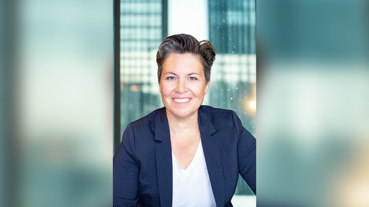 A portrait of a woman with short hair and wearing a dark blazer sitting indoors with her back to a window.