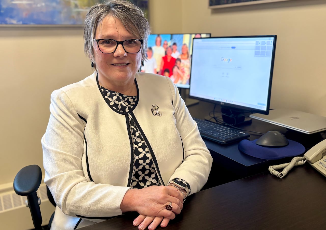 A woman in glasses and a white jacket sits near a computer with two screens.