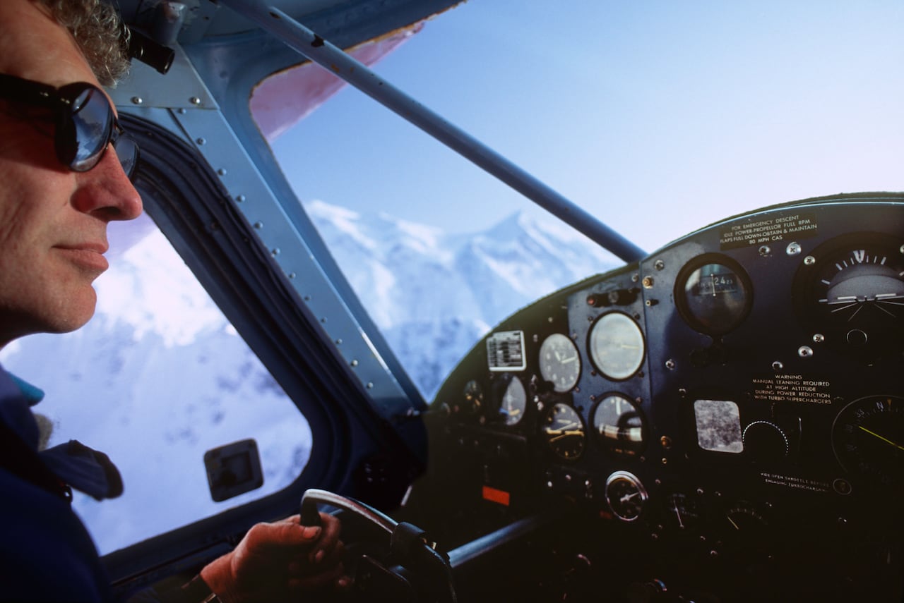 A pilot seen inside the cockpit of a small plane in the mountains.