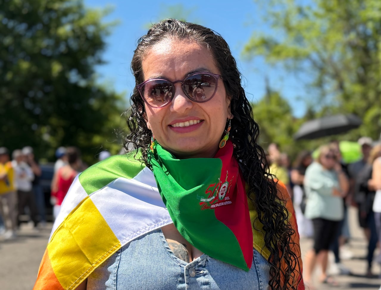 A woman smiling at the camera with a bandana around her neck and flag draped over her shoulders. She's outside and there's a line behind her