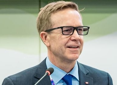 Portrait of a man with fair hair and dark glasses, wearing a blue suit.