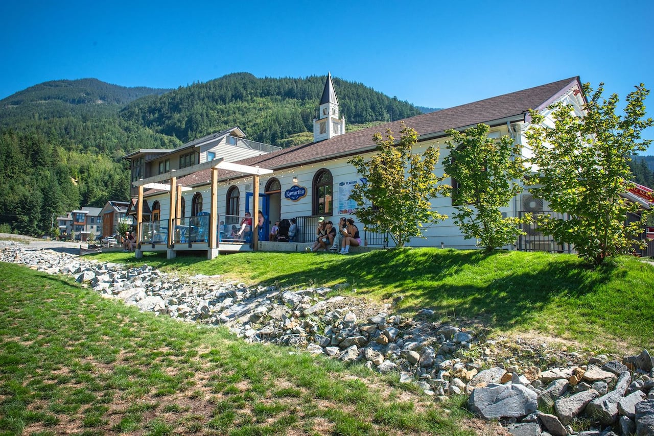 A white church-style building sits beside a grassy area with forested mountains behind it and creek running passing through it.