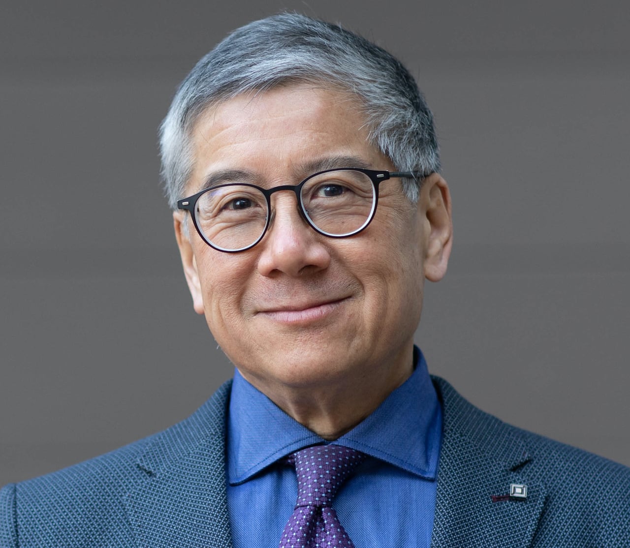 Portrait of a man with grey hair and glasses, wearing a blue suit, blue shirt and purple tie