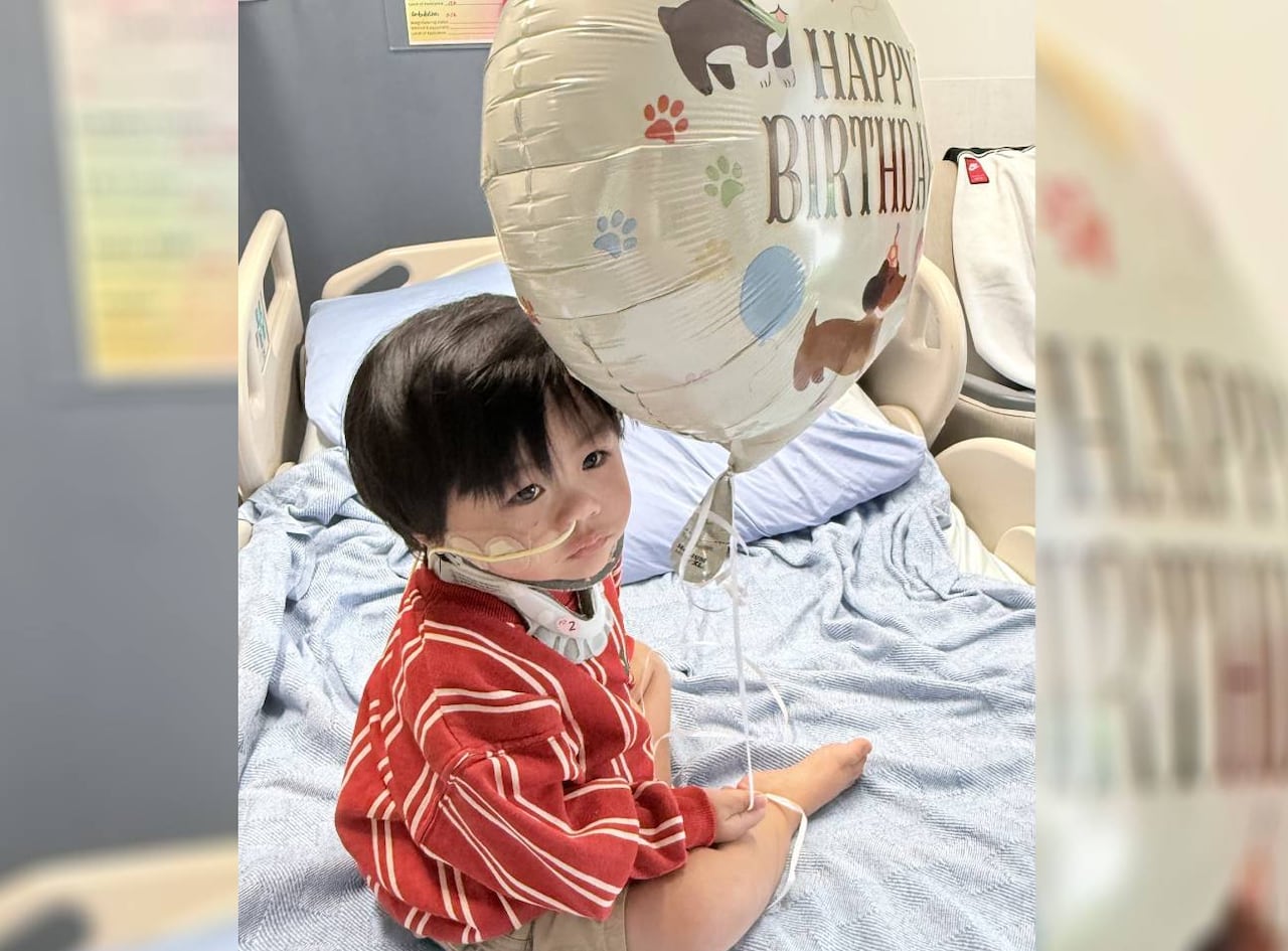 A toddler sit on a hospital bed and holds a balloon that reads, 'Happy Birthday.'