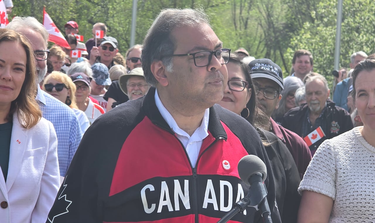 A man wearing glasses and a red, white and black jacket that says 'CANADA' on the chest stands at a podium. Supporters stand behind him. They are outside.