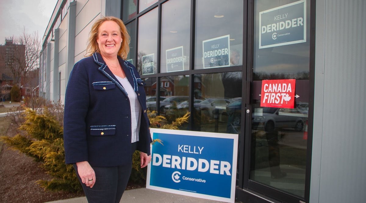 Woman in suit stands outside a building, there are blue Conservative signs that say her name "Kelly DeRidder."