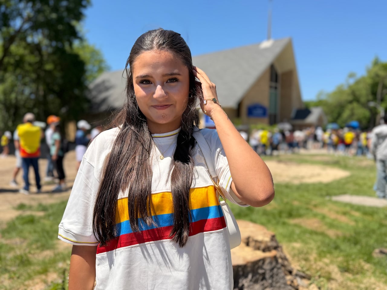 A girl smiles at the camera outdoors. She's wearing a shirt with the Colombian flag colours. There's a line of people behind her
