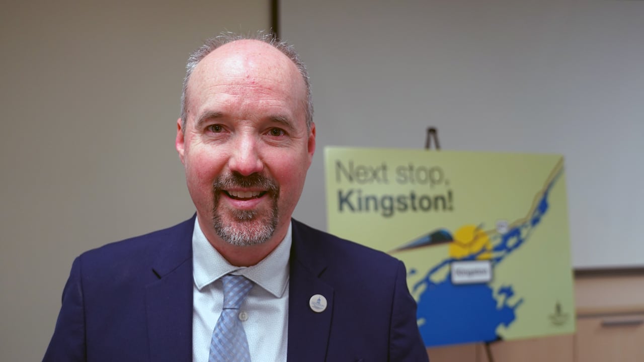 A man with short black and grey hair and a goatee stands in front of a sign that has a picture of a train and says "Next stop, Kingston!"