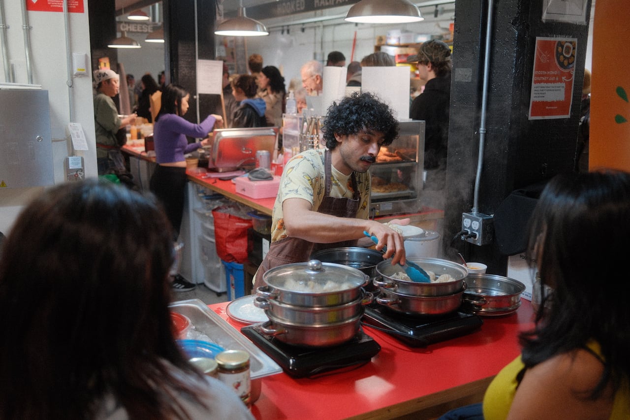 A young man with curly hair and a moustache is shown serving Nepalese dumplings at a farmers' market.