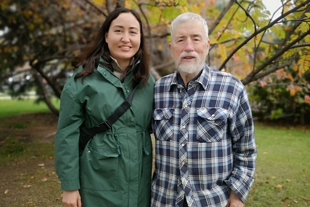 A woman in a green coat and an older man in a plaid button down stand side-by-side in a field.