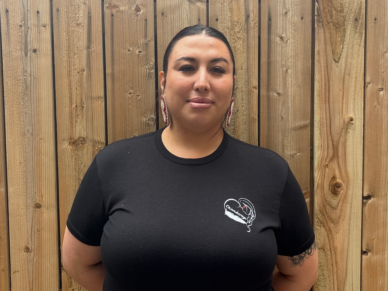 A woman with dark hair pulled back stands in front of a wooden fence, wearing a black T-shirt and beaded earrings.