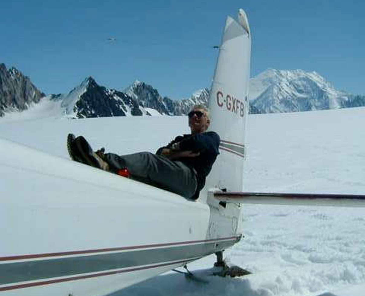 A man reclines on the tail of a small plane sitting among snowy mountains.