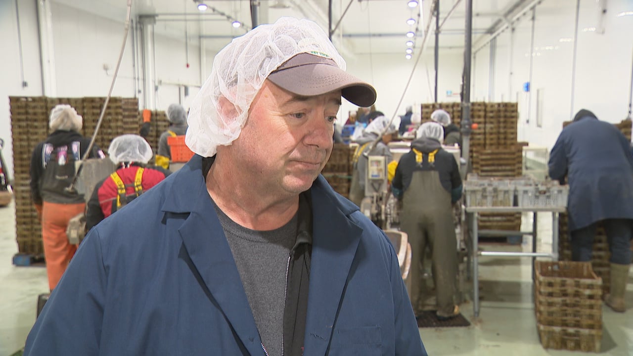 A man in a blue coat and wearing a hairnet over a hat stands in a factory.