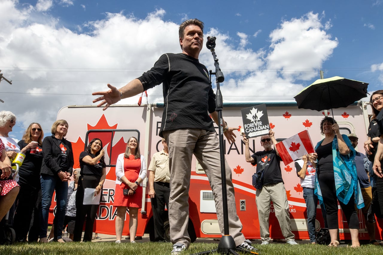 Man at a microphone spreads out his arms while talking in front of a red and white pro-Canada campaign bus.