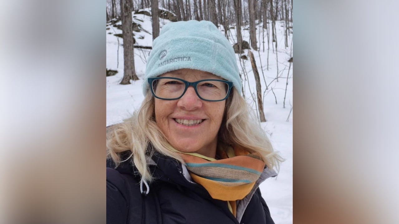 A woman with grey-blonde curly hair wears a powder blue tuque, a yellow and blue scarf and a dark jacket stands in a wooded area covered in snow,