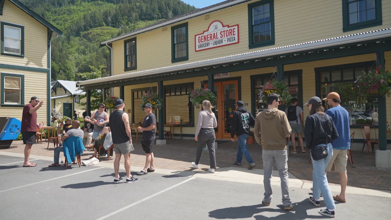 A group of people stand and sit outside a yellow general store building on a sunny day.