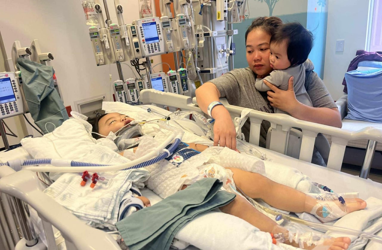 A toddler lays in a hospital bed, hooked up to various equipment, as a woman and baby look over him.