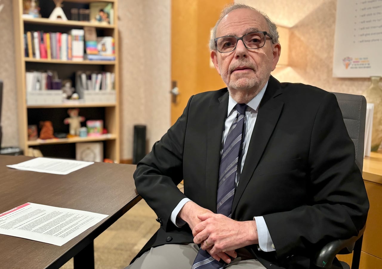 A man in glasses and a dark suit sits near a brown desk with a bookshelf in the background.