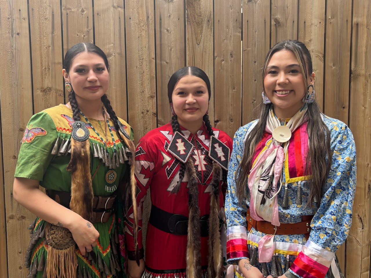 Three young women stand side by side wearing colourful jingle dresses.