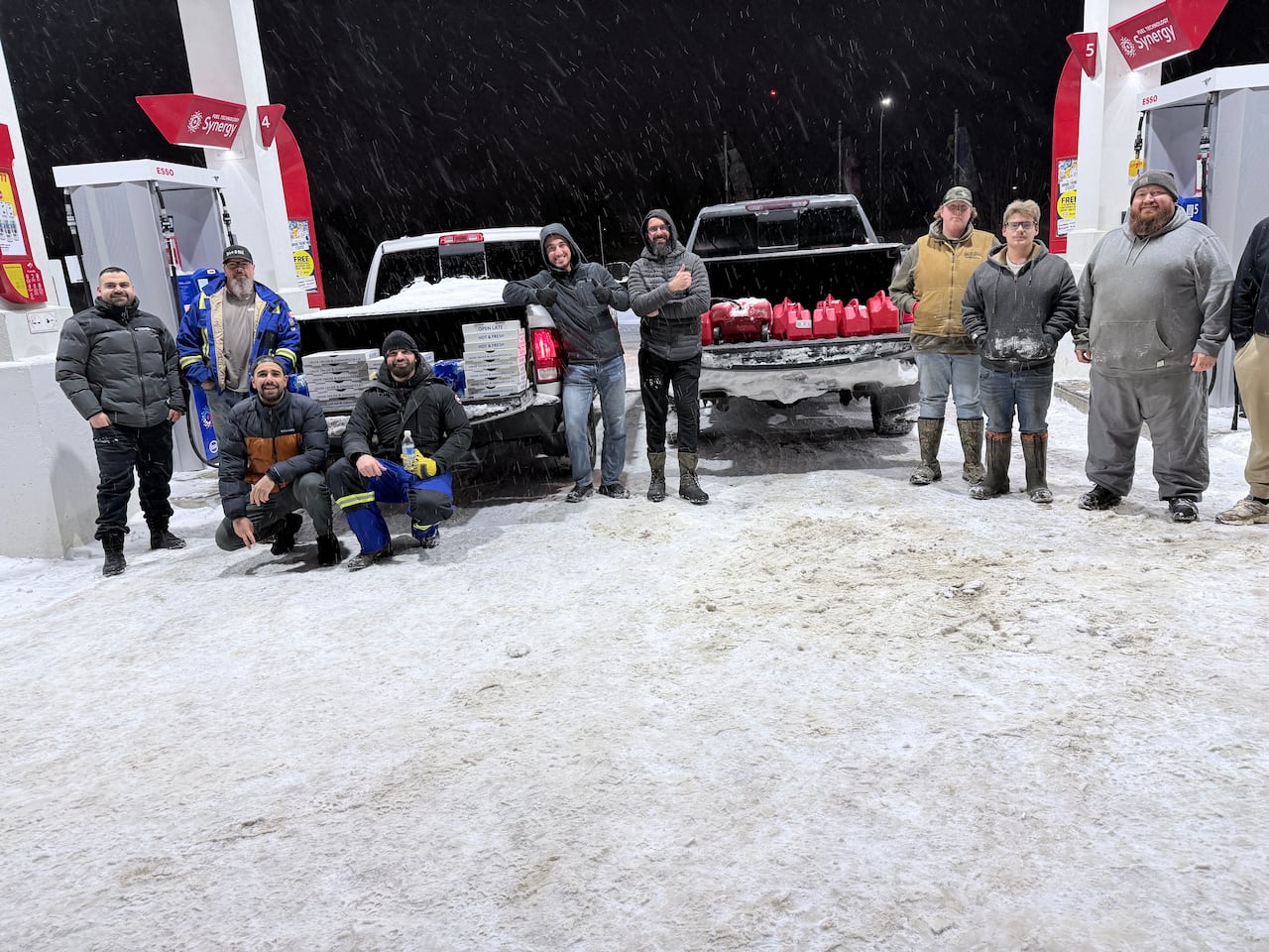 A bunch of men stand in front of a gas pump with two cars full of gas and food.