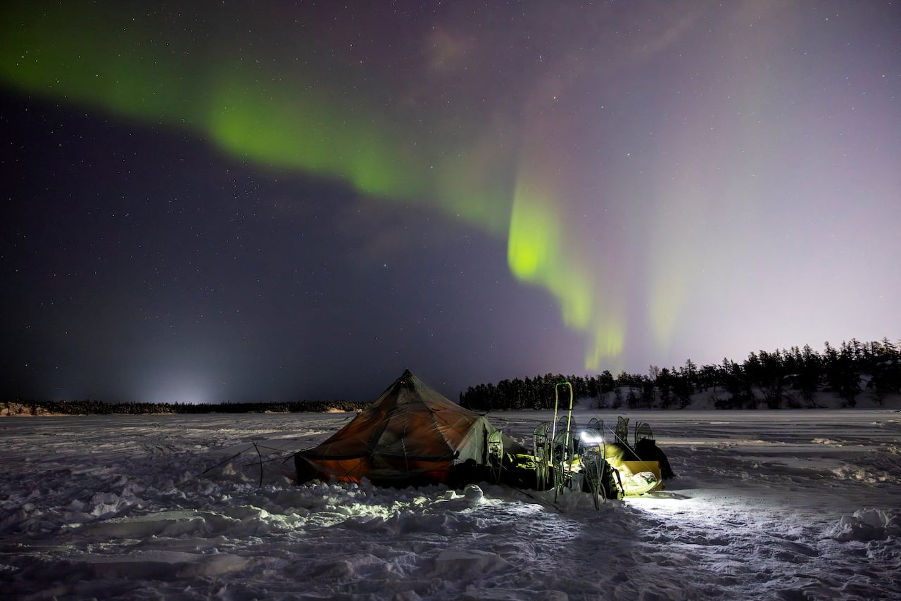 A tent on snow-covered barren land with northern lights dancing in the sky at night.