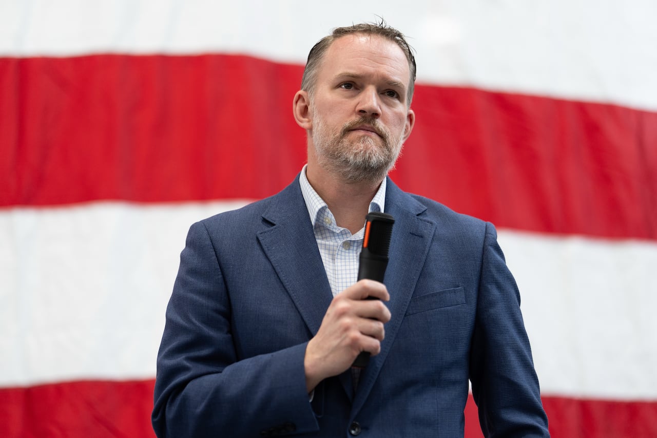 A man in a suit holds a microphone in front of a large American flag.