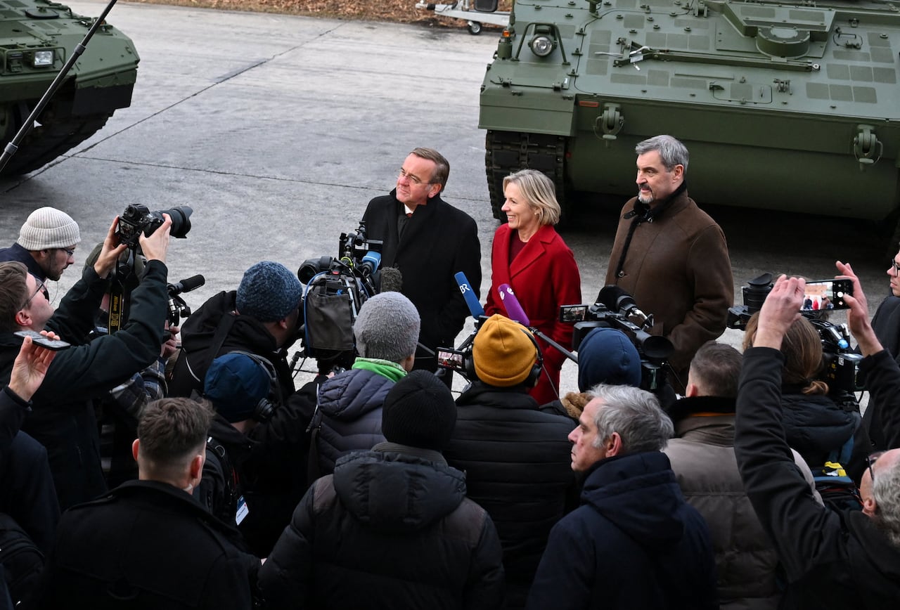A large group of reporters stand facing a group of politicians in front of a tank.