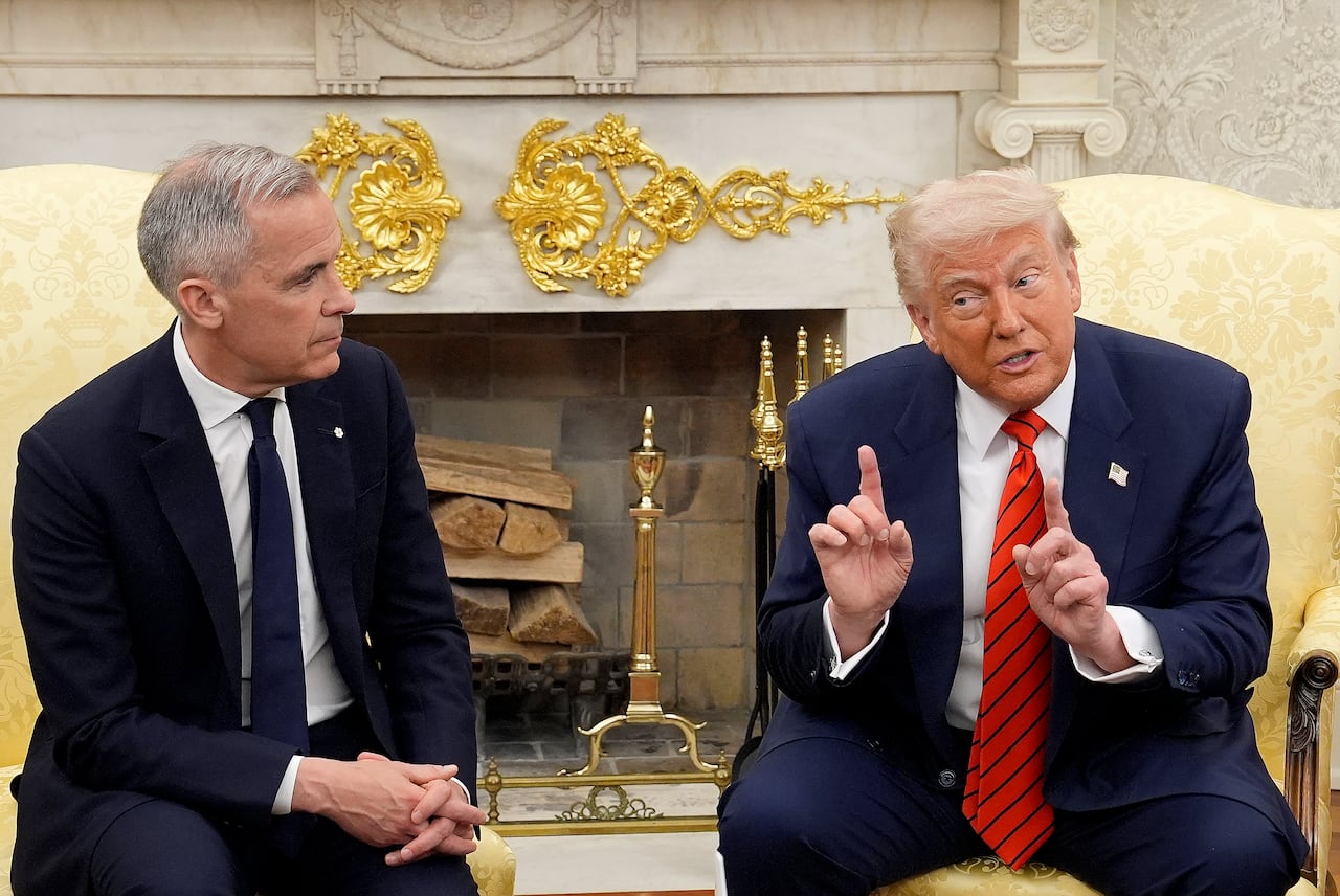 Two men in suits are seated in arm chairs in front of an ornate fireplace.