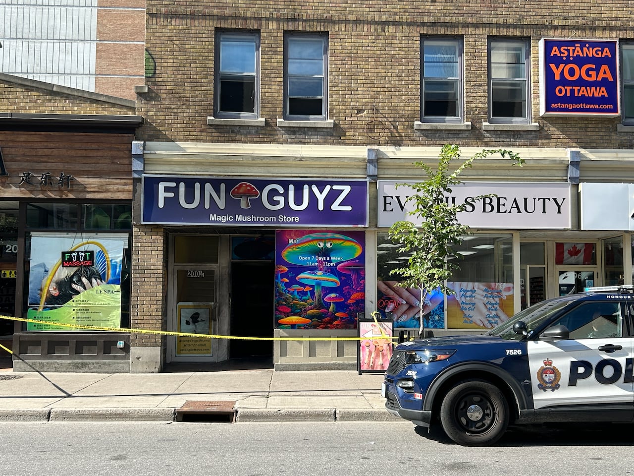 A police vehicle parked outside a magic mushroom store in a city's downtown. The store has yellow tape around it and its front is slightly blackened.