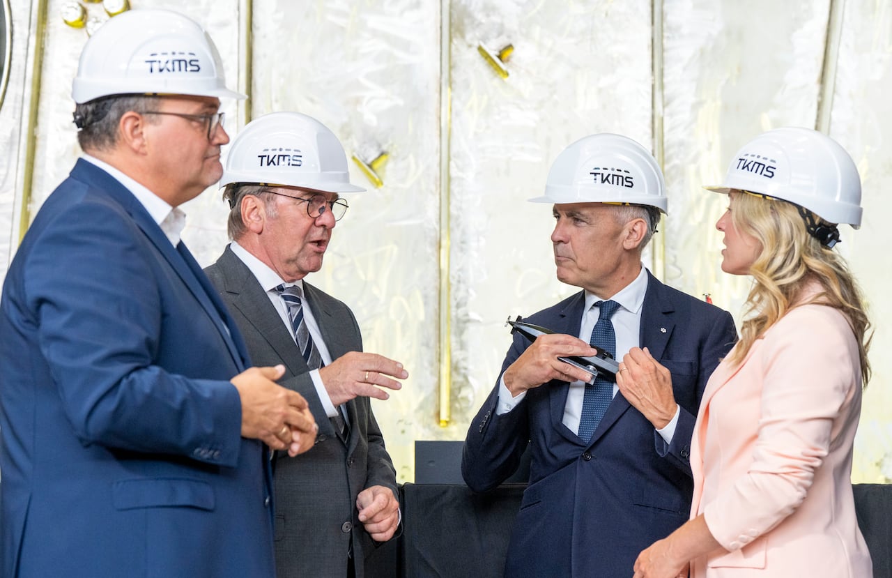 Three men and a woman wear suits and white hard hats as they stand together talking. The man second from right holds a model submarine and pretends to put it in his inner suit pocket.