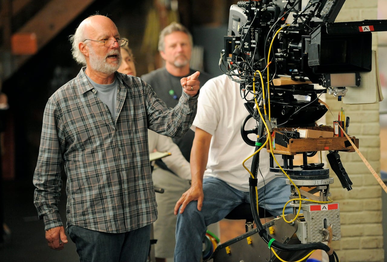 An older man with balding grey hair and glasses stands behind a television camera and points with his left hand as he directs a television show.