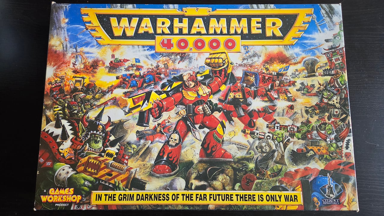 A cardboard box with art for a game is seen on a table. The art includes men wearing red armour firing big guns at green-skinned orcs.