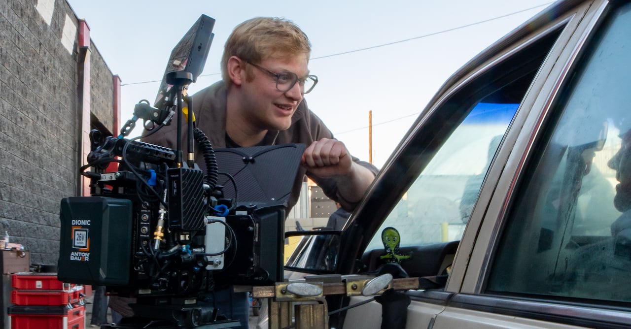 A man leans against a camera pointed into the driver's side of a parked car.