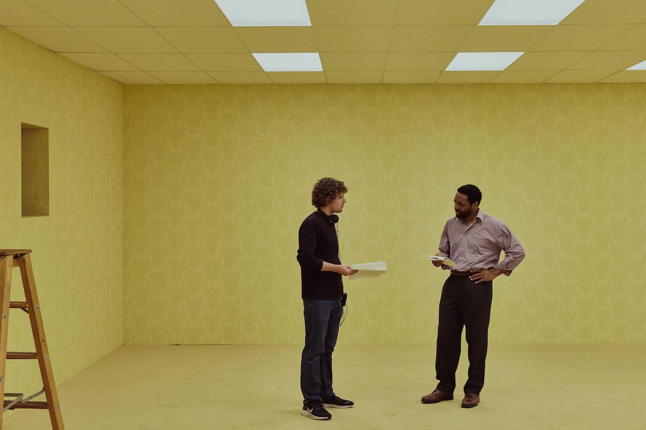 Two men stand, facing each other, in a large, yellow-ish room.