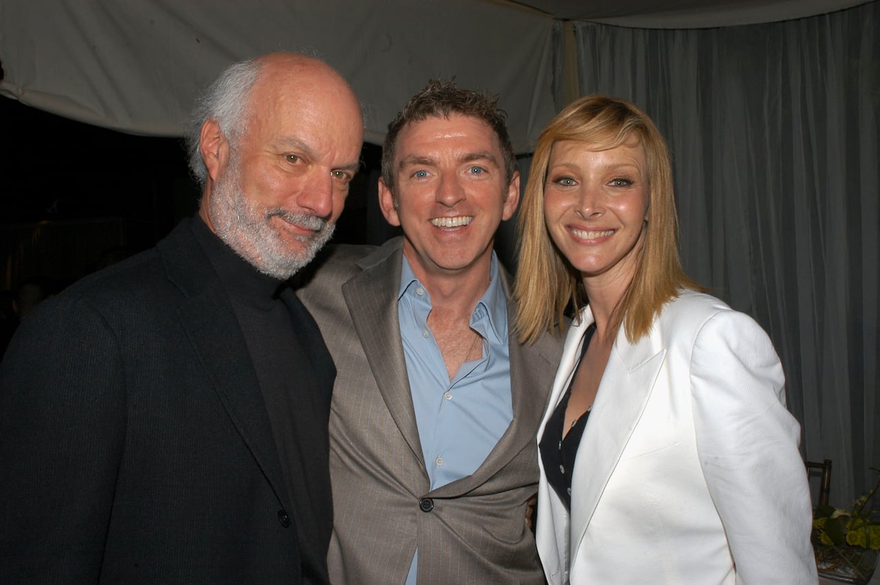 A balding, grey-haired man in black sweater stands next to a smiling younger man, wearing a light brown suit and light blue shirt, and a smiling blonde-haired woman in a white suit and black top.