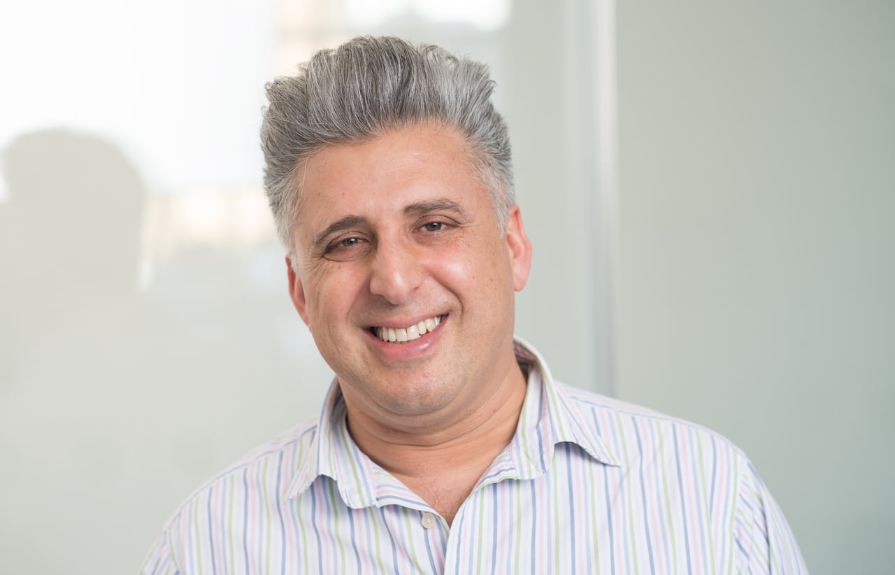 A man with grey hair and a striped, collared shirt smiles looking straight to camera.