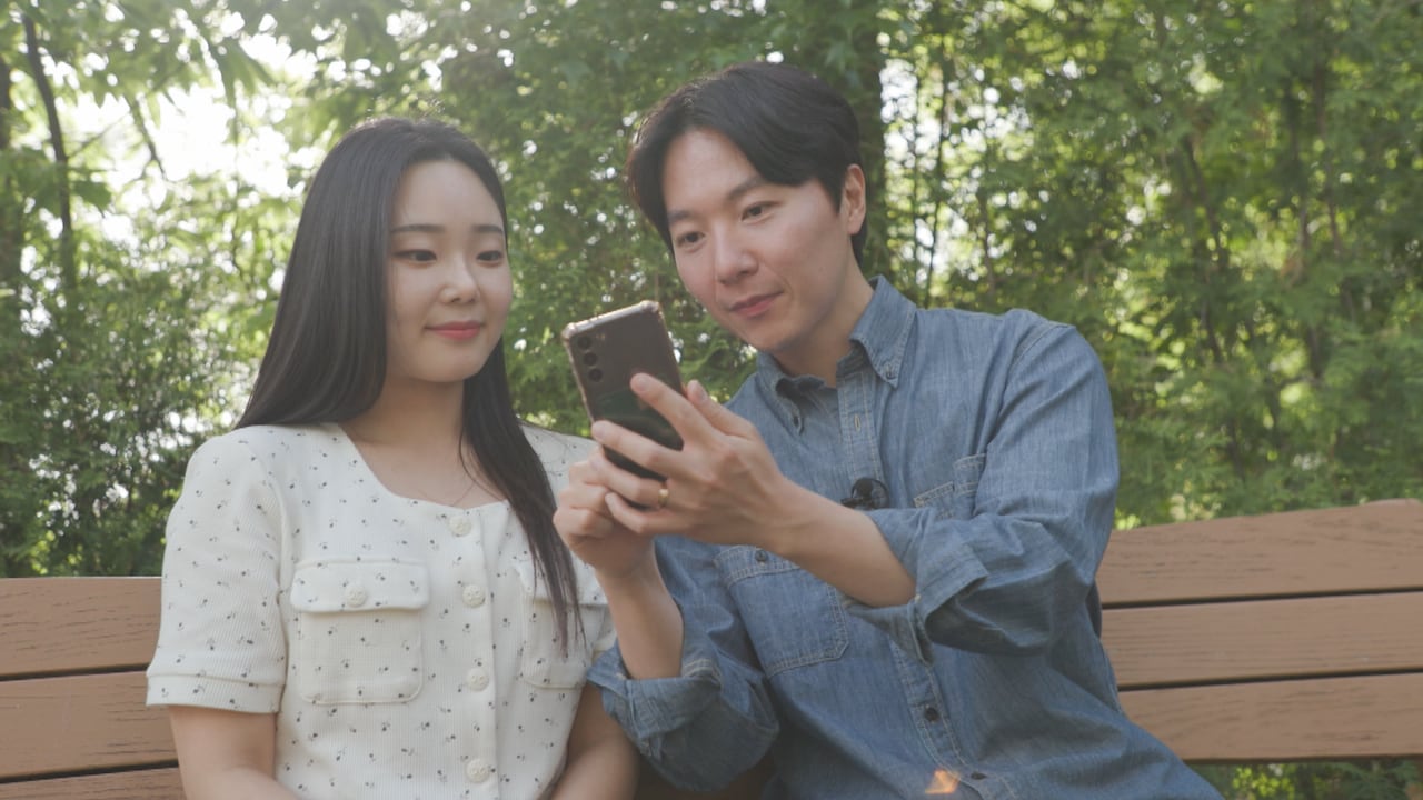A  couple sits on a park bench and smiles at a phone