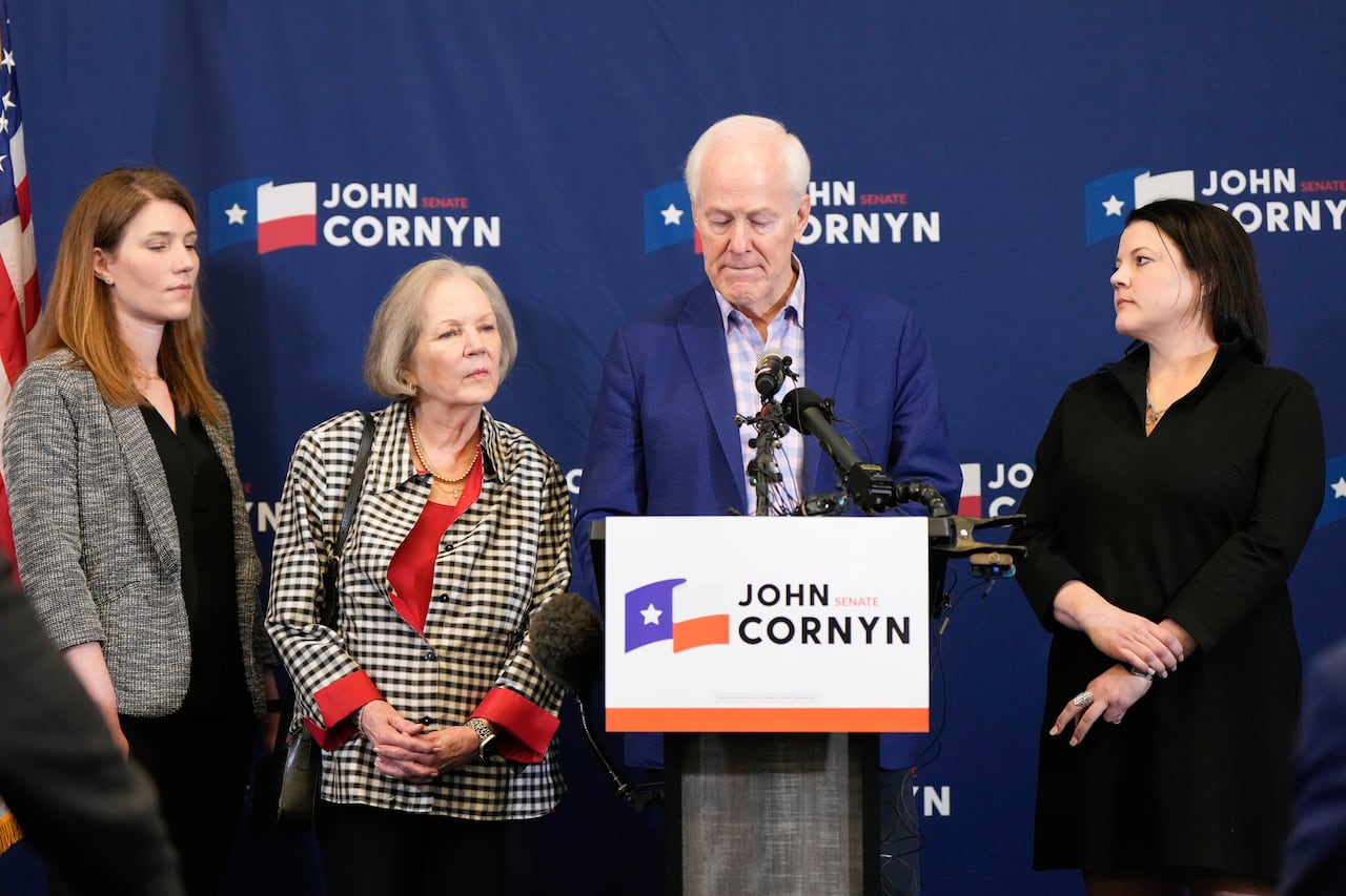 A white-haired man is shown standing behind a podium that says 'John Cornyn,' and is surrounded by three women of various ages.