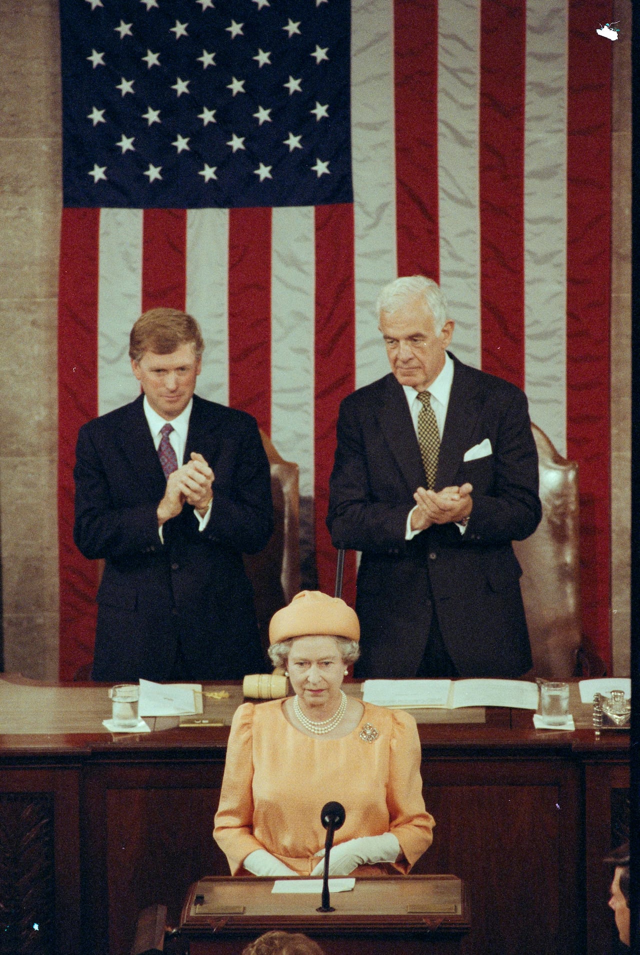 A person stands behind a microphone on a podium as two people standing behind applaud. A large American flag hangs vertically behind them.