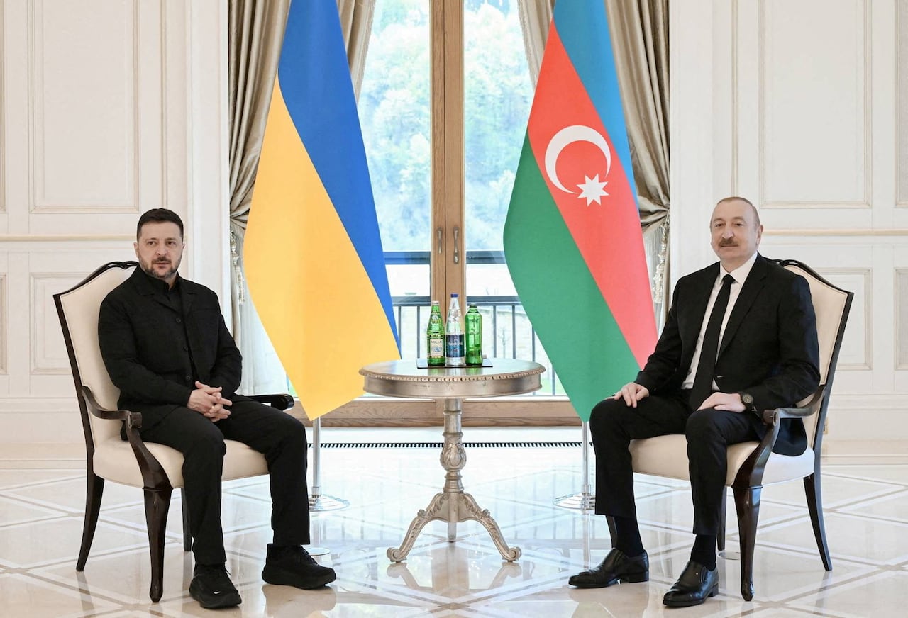 Two officials sit in chairs, positioned on either side of a table that is topped with bottled water. An Azerbaijan flag and a Ukrainian flag are placed between the two officials.