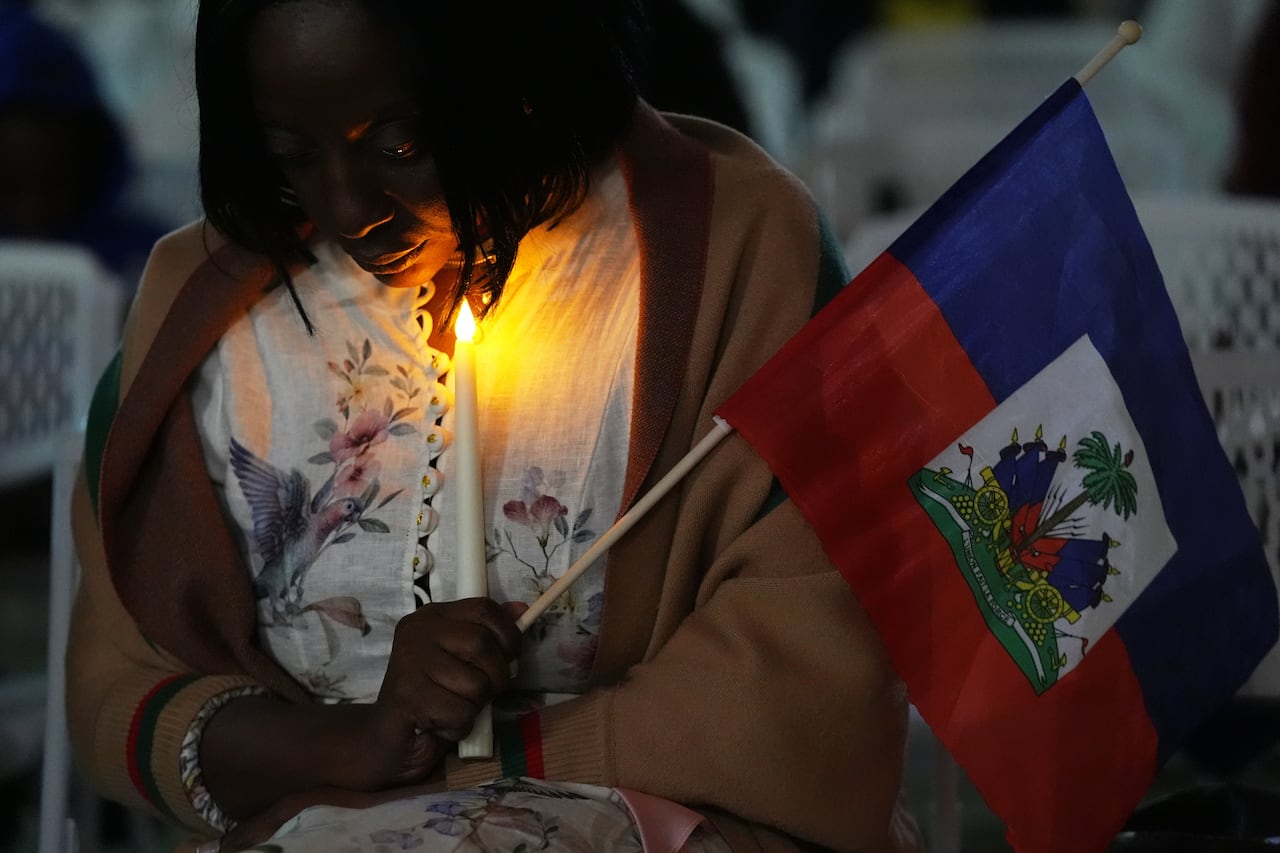A dark-complected woman holds both a candle and a small flag while bowing her head in a nighttime scene.