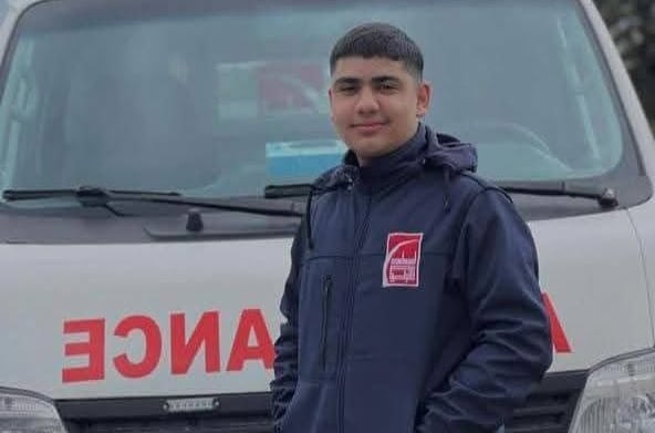 A smiling young man stands with his hands in the pockets of a dark jacket with a red patch as he stands in front of an ambulance.