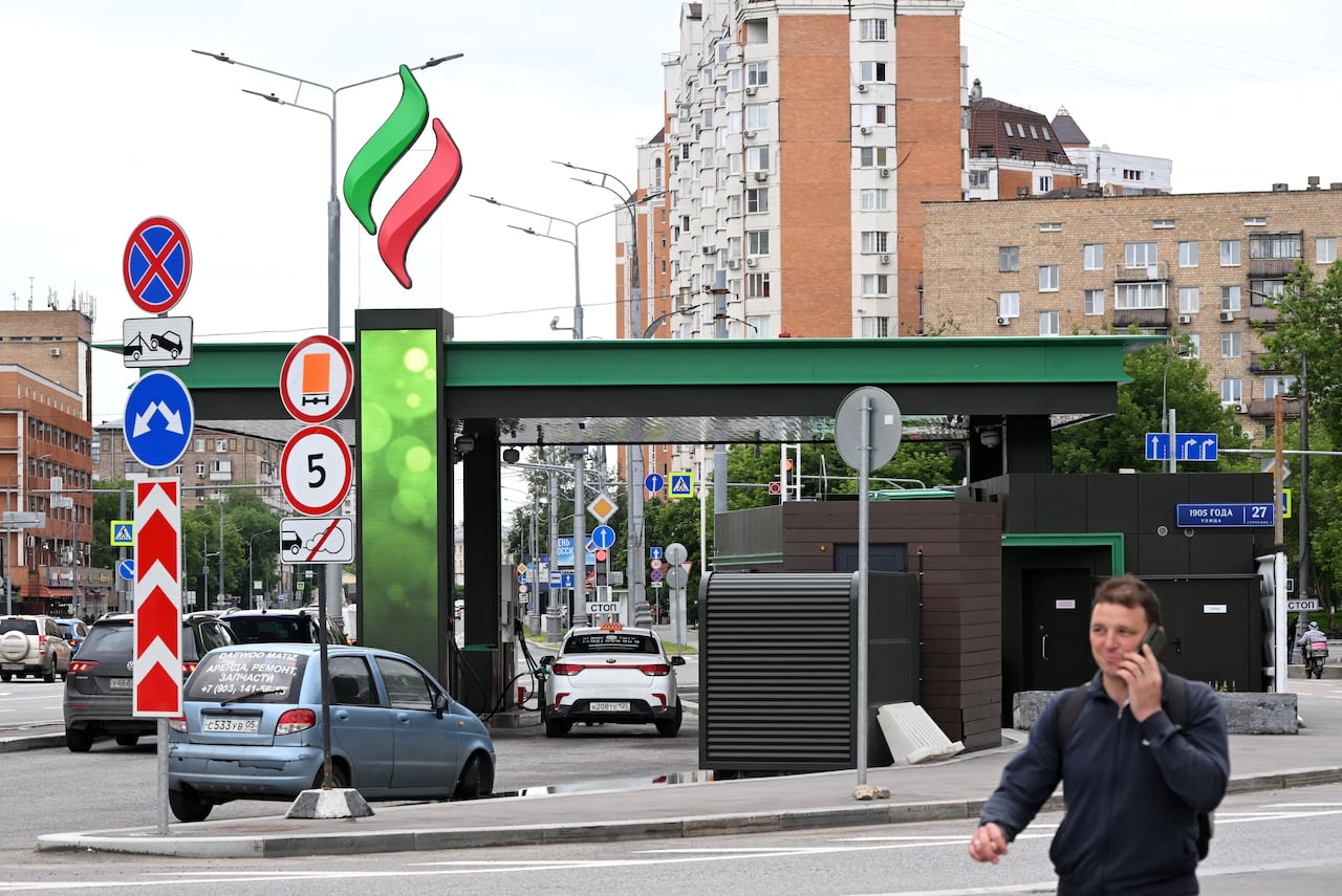 A person talks on a cellphone to their ear on a sidewalk as a gas station is shown near him.
