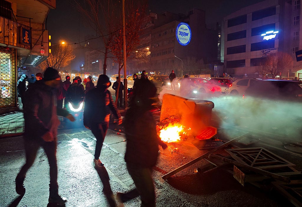 People walk by a burning object on a sidewalk.