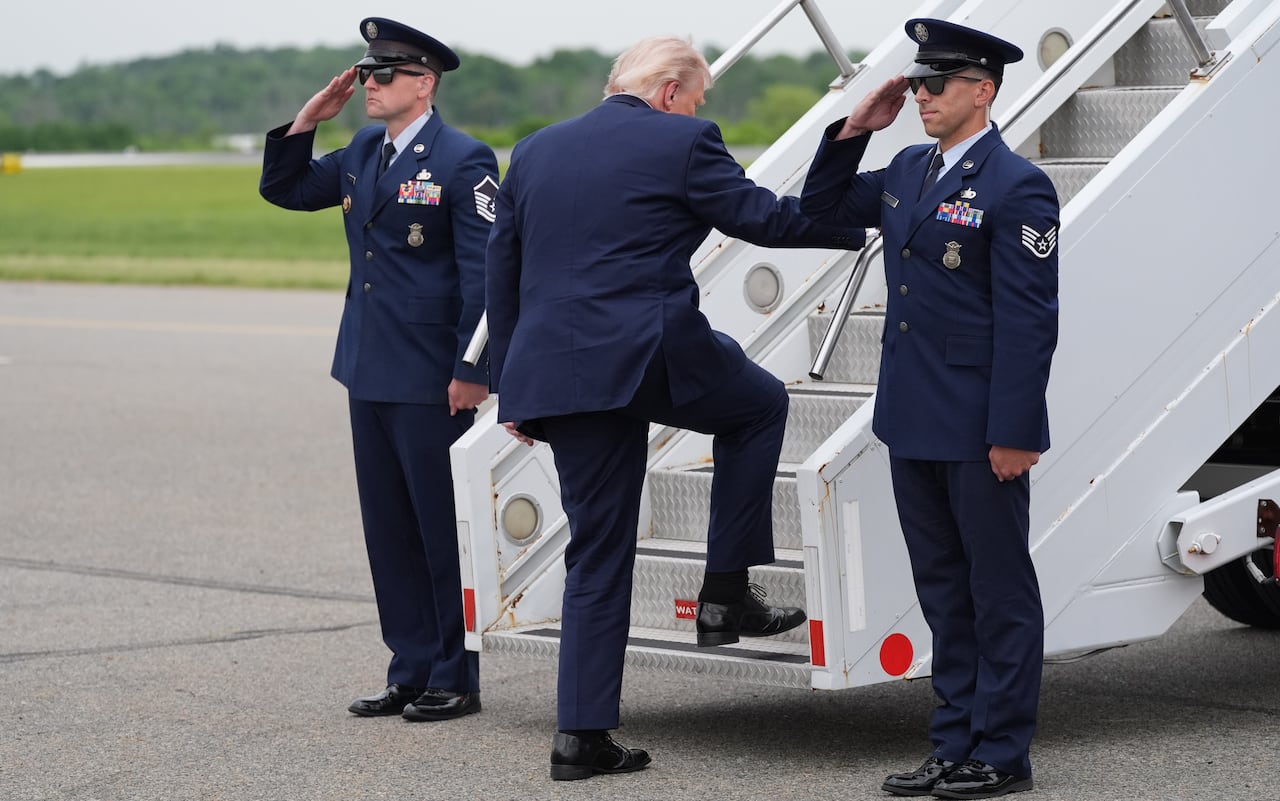 Flanked by two saluting marines, Donald Trump steps on the stairs leading to an airplane door.