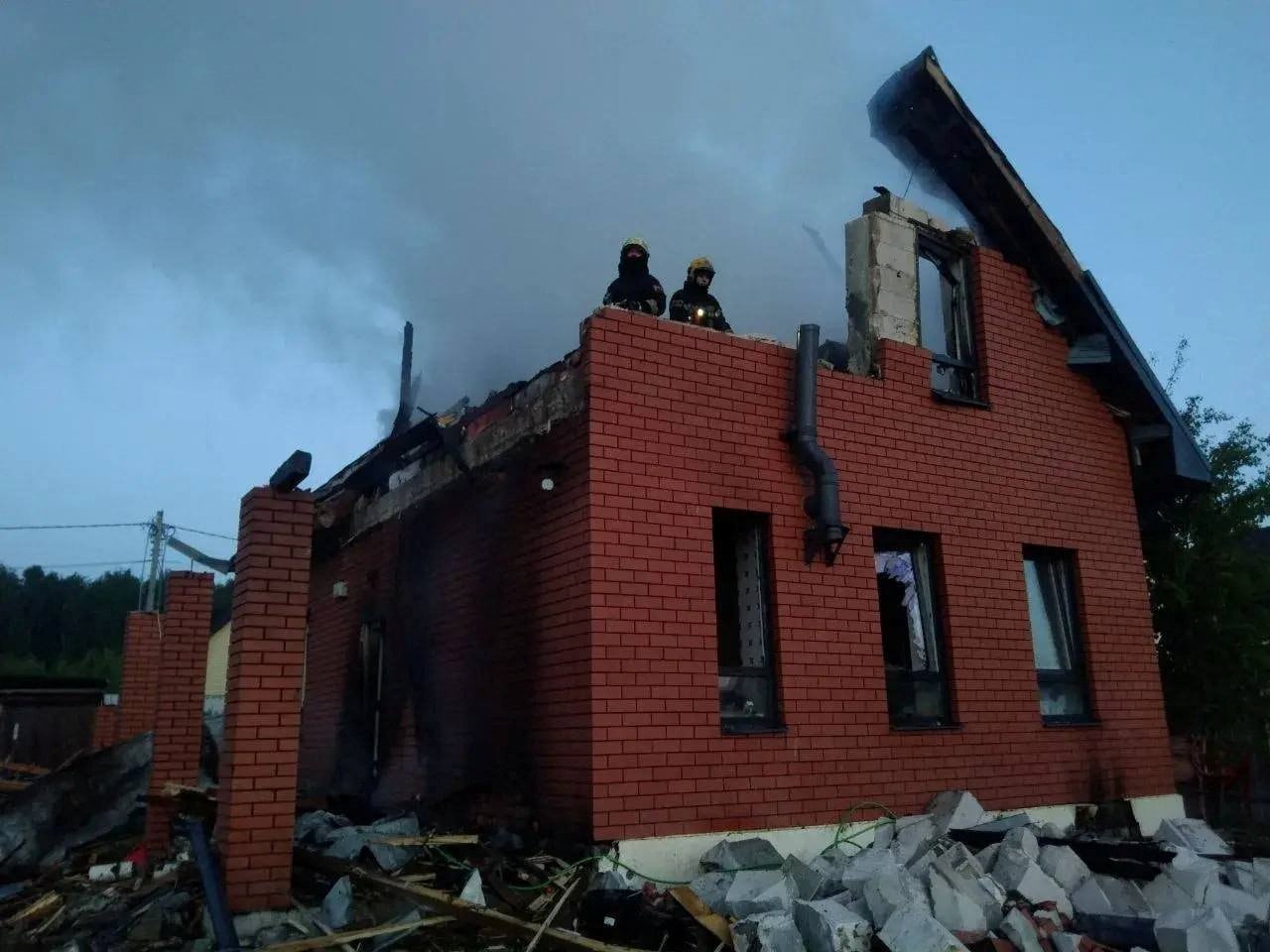 A small brick house is seen destroyed with no roof and two firefighters peering out from a top floor exposed to the sky.