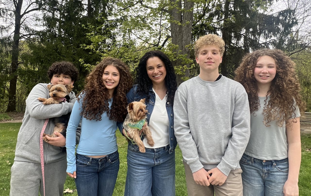 A woman holding a small dog smiles as she poses for a photo in a park surrounded by her four children, two boys (one of whom is also holding a small dog) and two girls.