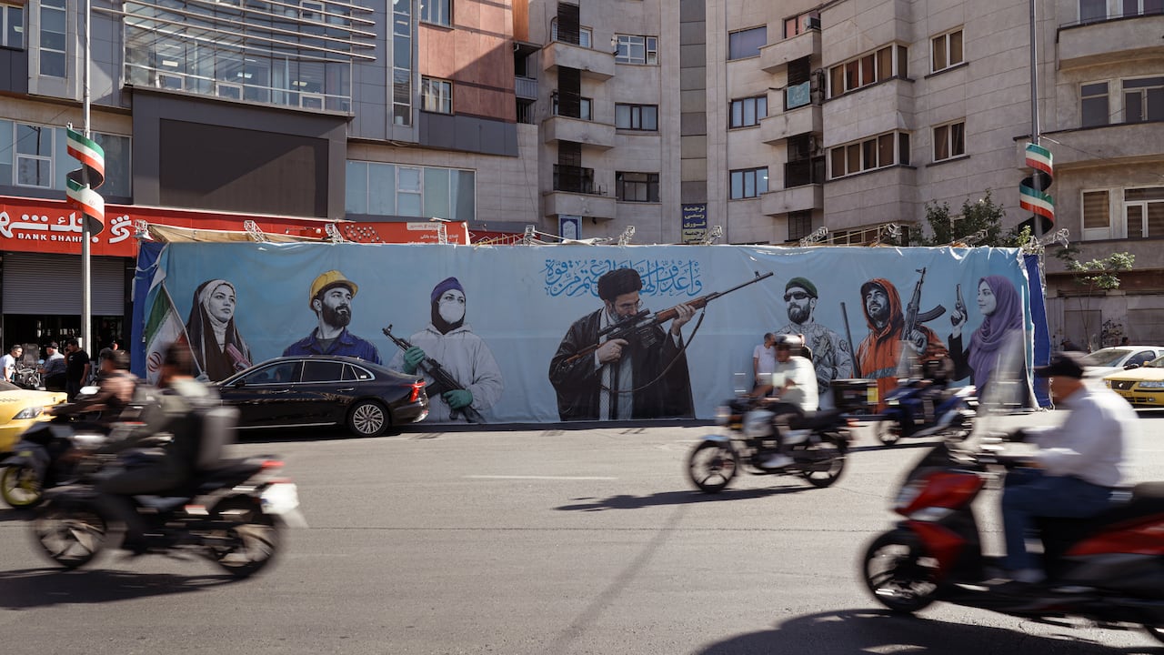 Motorcylces pass by a billboard with men holding guns displayed on it.